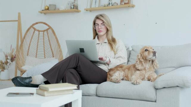 Young Woman in Glasses Using Laptop While Sitting on Couch with Her Dog. Background- Home Interior