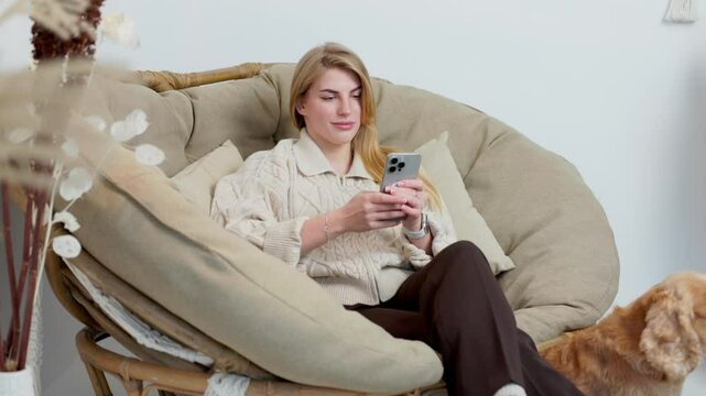 Young Woman Sits in Comfortable Wicker Chair with Her Pet Dog and Flipping Through Her Phone