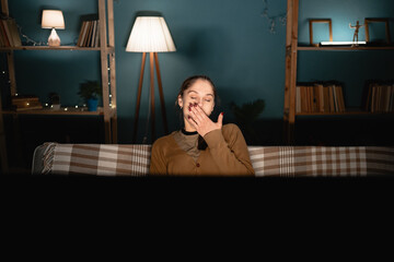 Young woman sitting on sofa in living room watching movie and yawning with tired expression.