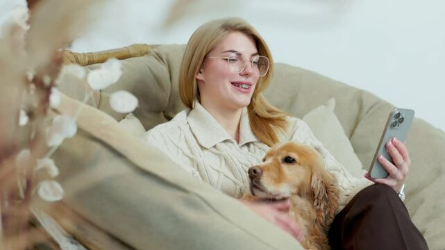 Young Woman Sits in Comfortable Wicker Chair with Her Pet Dog and Scroll Through Phone