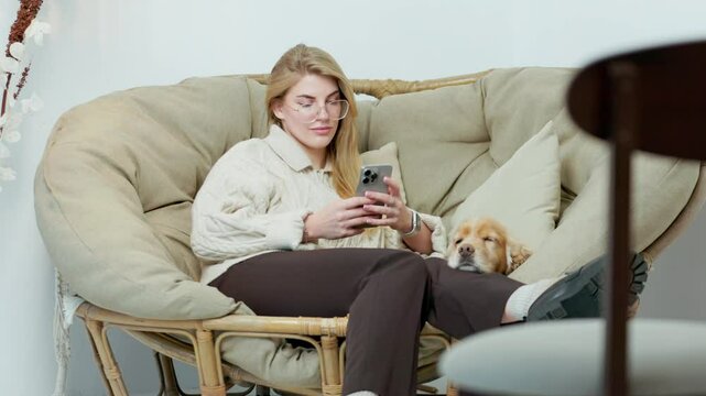 Young Woman Sits in Comfortable Wicker Chair with Her Pet Dog and Scroll Through Phone