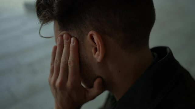 Close-up headshot of businessman experiencing intense discomfort from severe headache, pressing hands against temples, likely caused by stress or migraine, sitting on urban bench alone, slow motion.
