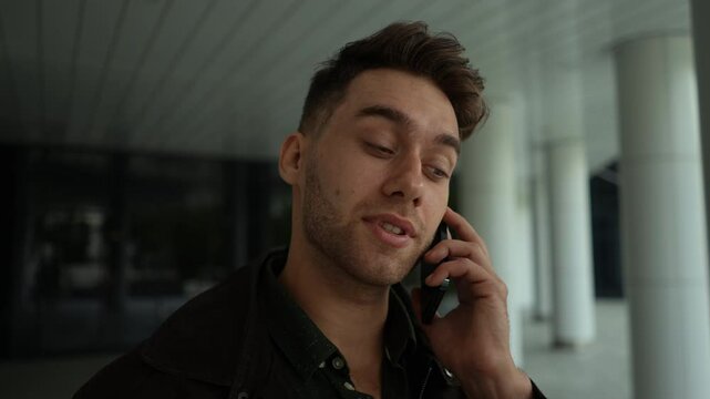 Portrait of focused young man having unpleasant conversation on mobile phone standing outdoors. Puzzled Caucasian male talking on smartphone with serious expression at urban street, slow motion.