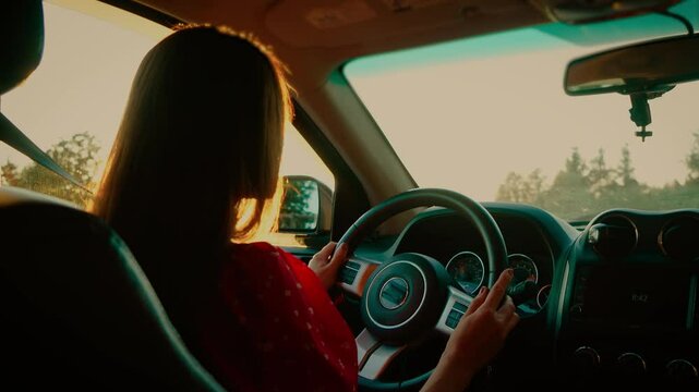 Young woman with straight hair driving car in red dress, holding steering wheel during sunset. Beautiful female focusing on road, evening light illuminating dashboard and interior. Calm lady steering