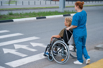 Obraz premium Red-haired nurse pushing an elderly woman in a wheelchair across the road. 