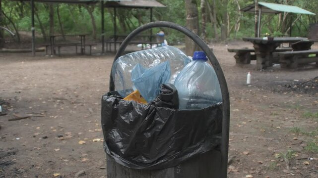 A trash bin in a park filled with rubbish. A dumpster near a public barbecue area.