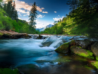 Obraz premium Serene mountain river flows over mossy rocks, lush green forests frame the vibrant scene under a dramatic sky. Sunlight illuminates the cascading water.