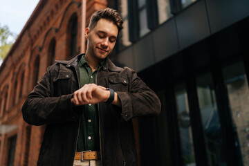 Confident successful businessman wearing brown suede jacket and green shirt smiling while checking time on smartwatch standing by office building, exuding punctuality and assurance.
