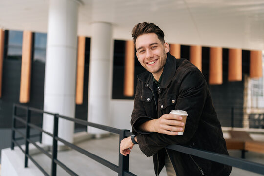 Portrait of happy young man holding takeaway coffee cup leaning on metal railing standing by modern building, smiling looking at camera. Serene dreaming male drinking enjoying coffee outdoors.