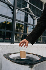 Close-up vertical cropped shot of unrecognizable busines man throwing disposable coffee cup into street trash can near office building, demonstrating responsible waste disposal and urban cleanliness.