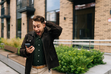 Happy excited young man standing in city street, holding mobile phone and reacting with astonishment to some unexpected news just received, looking to smartphone screen with amazed expression.