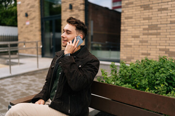 Handsome bearded young man having pleasant phone conversation while sitting on urban bench near modern building in urban environment, enjoying communication. Concept of successful city lifestyle.