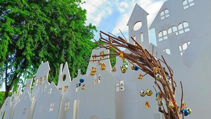 A charming artistic display showcasing a whimsical arch made of twigs, embellished with colorful ornaments, set against a backdrop of white, stylized houses and vibrant greenery