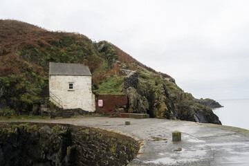 A calm coastal scene with a small fishing boat at low tide, a weathered building on a hillside, stone harbor structures, people strolling along the quay, and seabirds, all under a muted, overcast sky.
