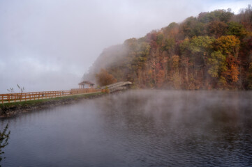 A serene lake is enveloped in mist, with a wooden walkway extending alongside it. Autumn foliage in vibrant reds and oranges covers the hillside, adding warmth to the cool, foggy atmosphere.