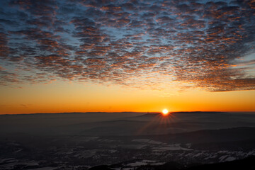 Golden sunrise over misty hills and snow-dusted valleys, painting the landscape in warm tones. A breathtaking view of peaceful nature under a dramatic, cloud-streaked sky.