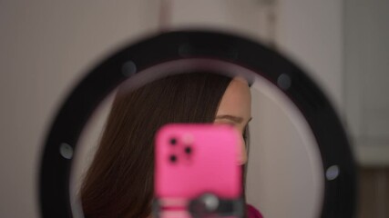 Young woman is talking to the camera, framed through a ring light with a pink smartphone in view, creating a modern and professional video setup. Concept of social media content creation and vlogging
