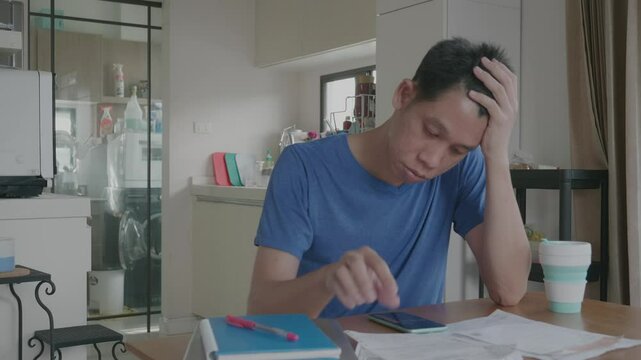 A man is sitting at a table with a cell phone and a stack of papers. He is focused on the papers and is not looking at the phone