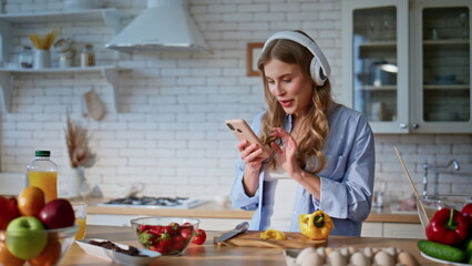 Joyful woman using mobile phone in kitchen while cooking. Lady listening music