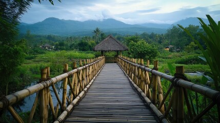 Obraz premium Bamboo bridge leading to lush green landscape with mountains in the background and a traditional bungalow under a cloudy sky.