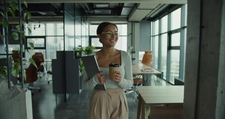 Modern professional woman with glasses holding a coffee cup and laptop walking confidently in a bright office space with panoramic city views and contemporary interior design - Powered by Adobe
