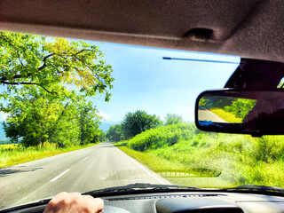 Car salon, windshield, hand of woman on steering wheel and landscape. View from seat of driver on nature with Road, trees, blue sky at sunny day