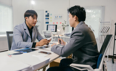 Two businessmen are sitting at an office desk, examining financial charts, data reports, and having a discussion about business strategies