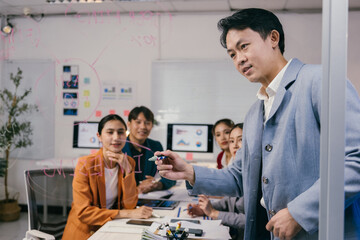 Asian manager drawing business plan on transparent whiteboard using pink marker during presentation to colleagues in office meeting room