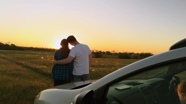 Married family couple in love hugging at sunset by car, romantically spending quiet time together, relaxing outside car. Husband and wife, spouses hugging, standing in field by car. Road trip, travel