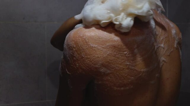 Slender woman with problem skin on her back applies soapy foam with washcloth to her shoulder in shower