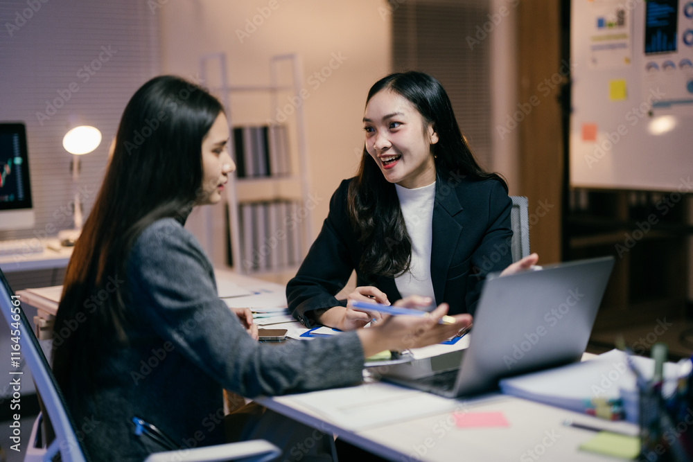Poster two young asian businesswomen are working late in the office, sitting at a desk and discussing a pro
