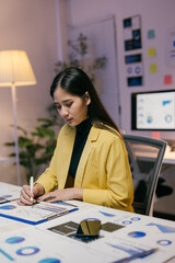 Young asian businesswoman concentrating on analyzing financial charts and graphs using pen and...