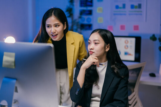 Two young businesswomen are working late in the office, collaborating on a project and analyzing data on a computer screen, demonstrating teamwork and dedication