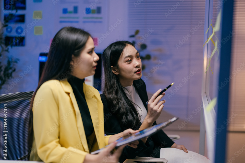 Poster two young businesswomen are working late in the office, brainstorming ideas using sticky notes on a 