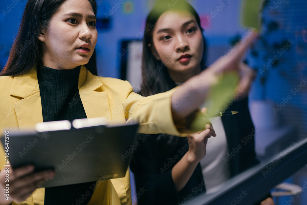 Poster two businesswomen are discussing and planning their strategy using sticky notes on a glass wall in a