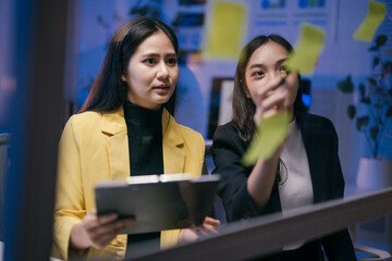 Two young businesswomen are using sticky notes on a glass wall to plan their business strategy, working late at night in a modern office