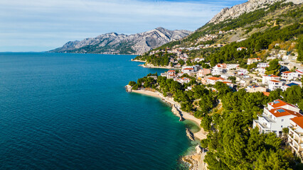 A stunning aerial view of Brela, Croatia, showcasing the iconic Kamen Brela rock formation surrounded by crystal-clear turquoise waters. The coastline is dotted with pristine beaches