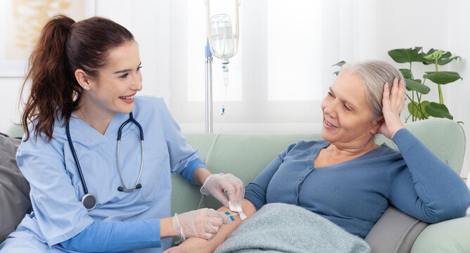 Young smiling nurse or doctor help sick senior elderly woman patient in receiving IV drip infusion and vitamin injection therapy sitting on sofa in medical clinic or at home. Health care concept