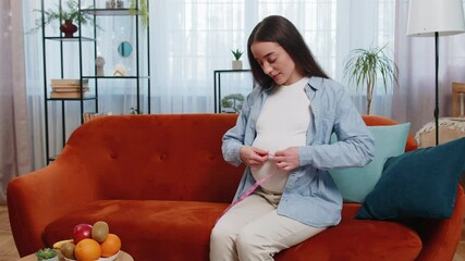 Smiling pregnant woman on sofa measuring belly with measure tape. Young future mother lady expecting awaiting childbirth monitoring body changes weight gain dimensions. Pregnancy maternity concept.