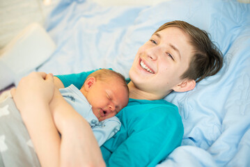 12 year old boy hugs his sleeping newborn brother. Happy teenager lies with his baby brother on the bed.