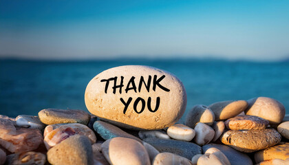 A smooth beach stone with "Thank You" written on it sits among natural pebbles against a blue ocean backdrop.