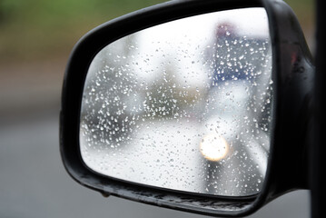 rear view mirror of black car, car is driving on road, there are a lot of raindrops on mirror, it is raining, road and car moving from behind are reflected in mirror