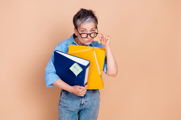 Elegant elderly woman holding colorful binders in casual outfit on beige background. She's adjusting her glasses