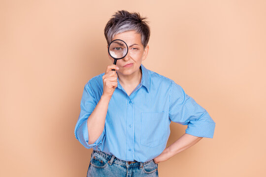 Elderly woman in stylish casual outfit holds magnifying glass with a curious expression on beige background