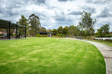 A well-maintained park featuring a green lawn, a paved walking path, trees, and a covered seating...