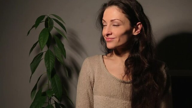 Happy carefree enjoing rest indoors woman with cute smile looking happy and relaxing sits on sofa at home interioor near flower in studio light, low key, dark shadow. Emotional people closeup video