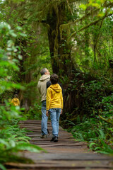Obraz premium Father with two kids are walking on a boardwalk in the rainforest with dense green foliage, ferns and cedars. Big tree trail, Vancouver island, British Columbia, Canada.
