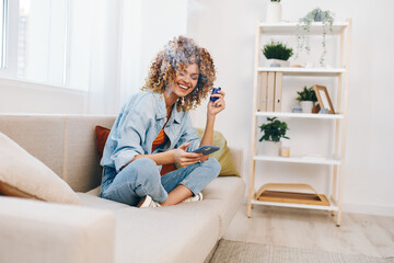 Cheerful woman sitting on a cozy sofa at home, holding a smartphone and smiling while chatting online, enjoying leisure time in her beautiful living room.