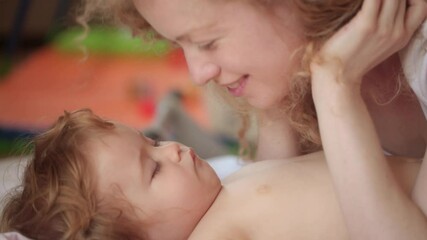 Happy young mother with little baby child lying on bed at bedroom. Beautiful mummy kissing and hug sleeping baby. Mother kissing sleeping baby. Mother caring for lovely child.