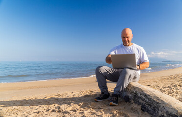 An adult Caucasian man, sitting on a log on the beach, working with a laptop
